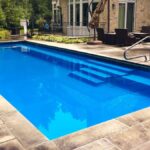 A backyard with a rectangular fiberglass pool surrounded by stone pavers, outdoor seating, and greenery.