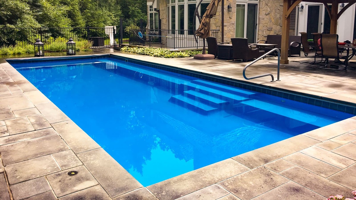 A backyard with a rectangular fiberglass pool surrounded by stone pavers, outdoor seating, and greenery.